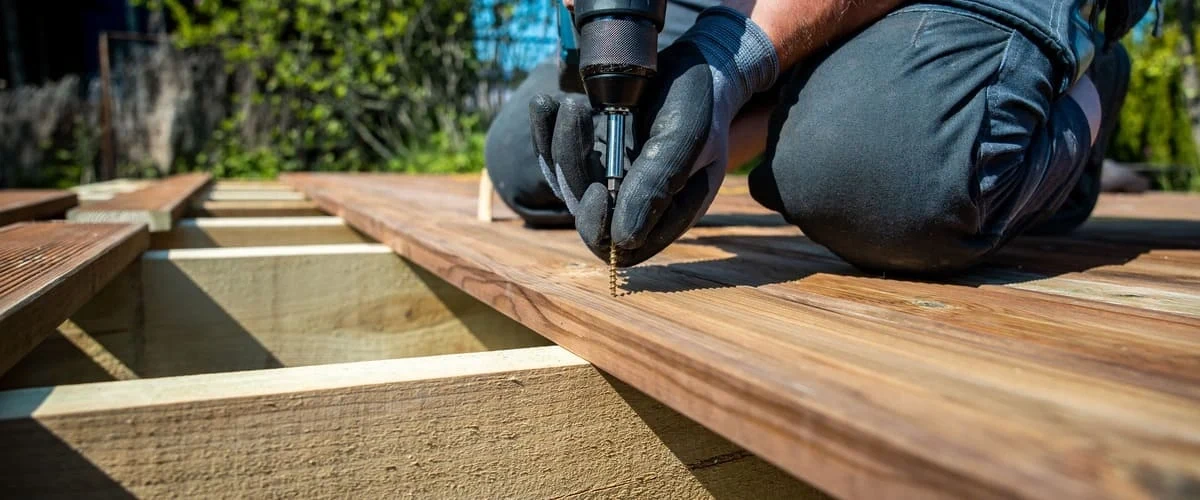 A close-up view of a professional contractor wearing safety gloves while using a power drill to drive a screw into wooden planks during a residential deck installation project.