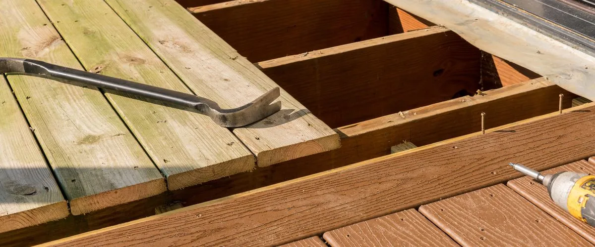 A close-up view of an outdoor wooden deck repair in progress, showing a crowbar prying up old light-colored wood planks to reveal the structural joists alongside new brown composite decking and a power drill.