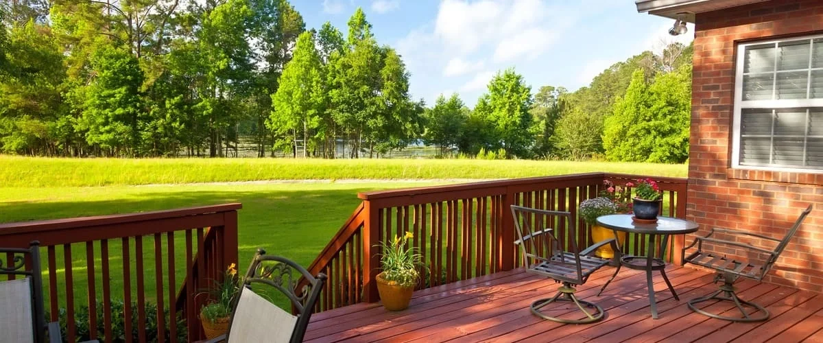 A spacious red-stained wooden deck featuring a metal patio dining set with swivel chairs, a glass-top table, and potted flowers, overlooking a lush green lawn and a dense line of trees under a clear blue sky.