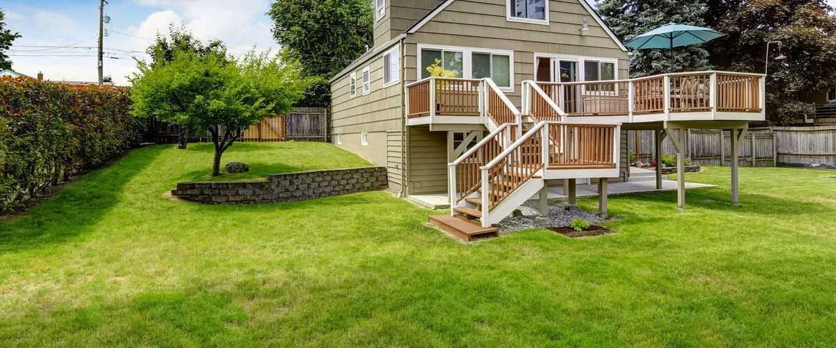 A high-angle view of a multi-level wooden deck attached to a two-story tan house, featuring sturdy support posts, white railings with brown balusters, and a clean staircase leading down to a green manicured lawn.