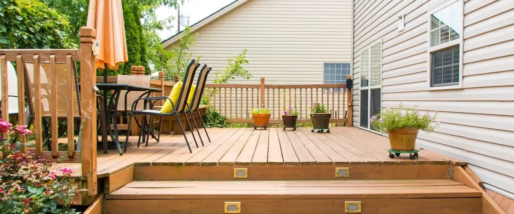 medium-sized outdoor wooden deck attached to a house with light beige siding. The deck features a dining set with an orange umbrella, black metal chairs with yellow cushions, and several potted flowers on rolling stands.