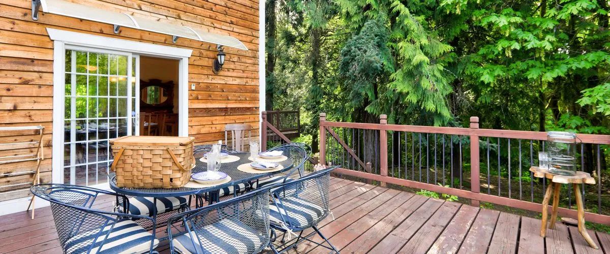 A spacious wooden outdoor deck with a black mesh dining table set for four, featuring a large picnic basket and striped cushions, overlooking a lush green mountain forest.