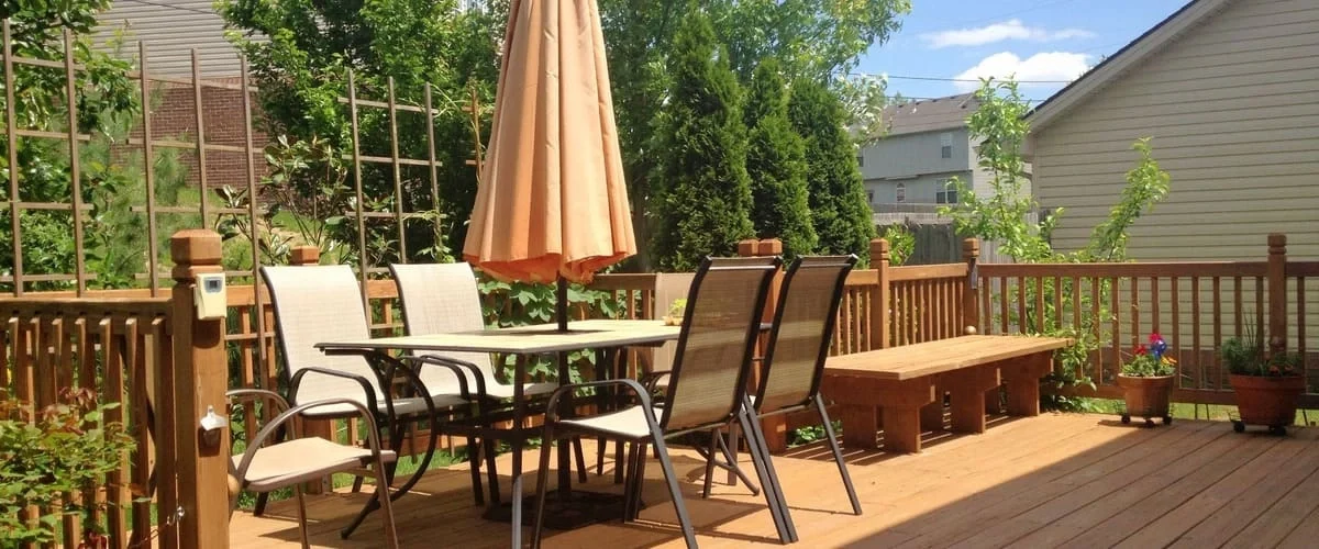 A spacious wooden outdoor deck featuring a rectangular dining table with four mesh chairs, a closed beige patio umbrella, a built-in wooden bench, and potted plants under a clear blue sky.