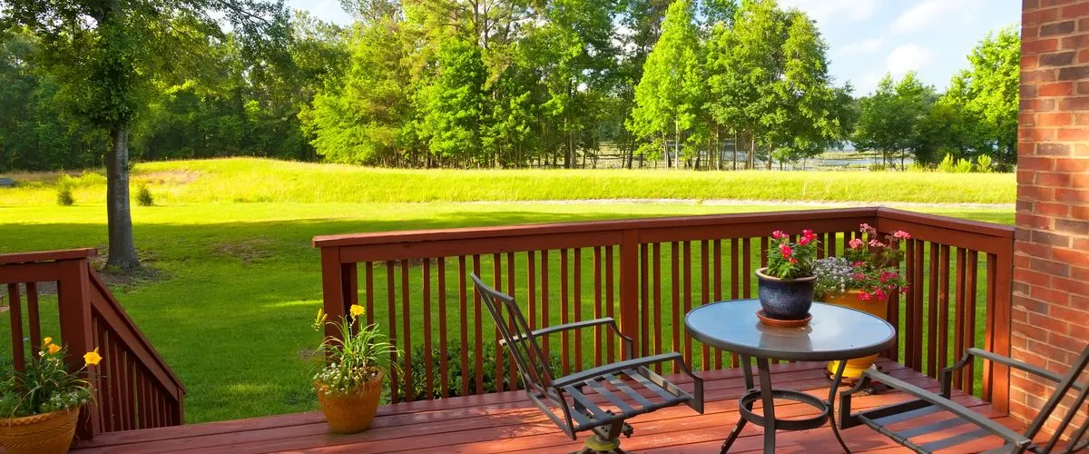 A scenic view from a redwood-stained wooden deck featuring a round glass-top table, metal chairs, and vibrant potted flowers, looking out over a sunlit grassy field and a dense line of green trees.