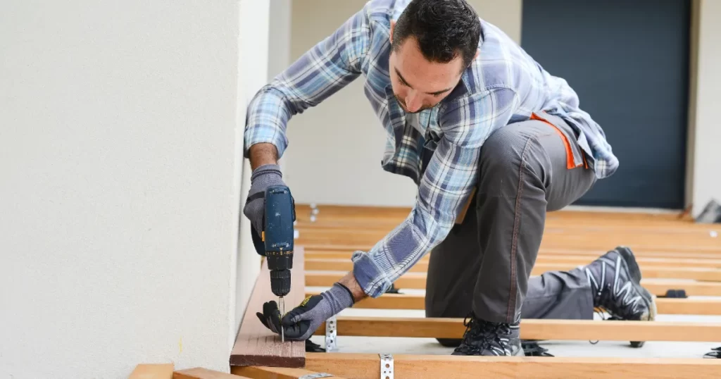 A carpenter is shown kneeling and using a cordless drill to secure a wooden plank to floor joists