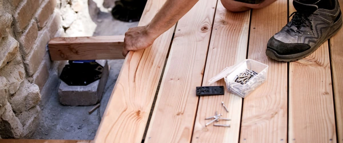 A close-up view of a professional deck installation featuring a worker’s hand aligning a light-colored wooden plank next to a stone wall, with screws and tools visible on the newly laid timber surface.