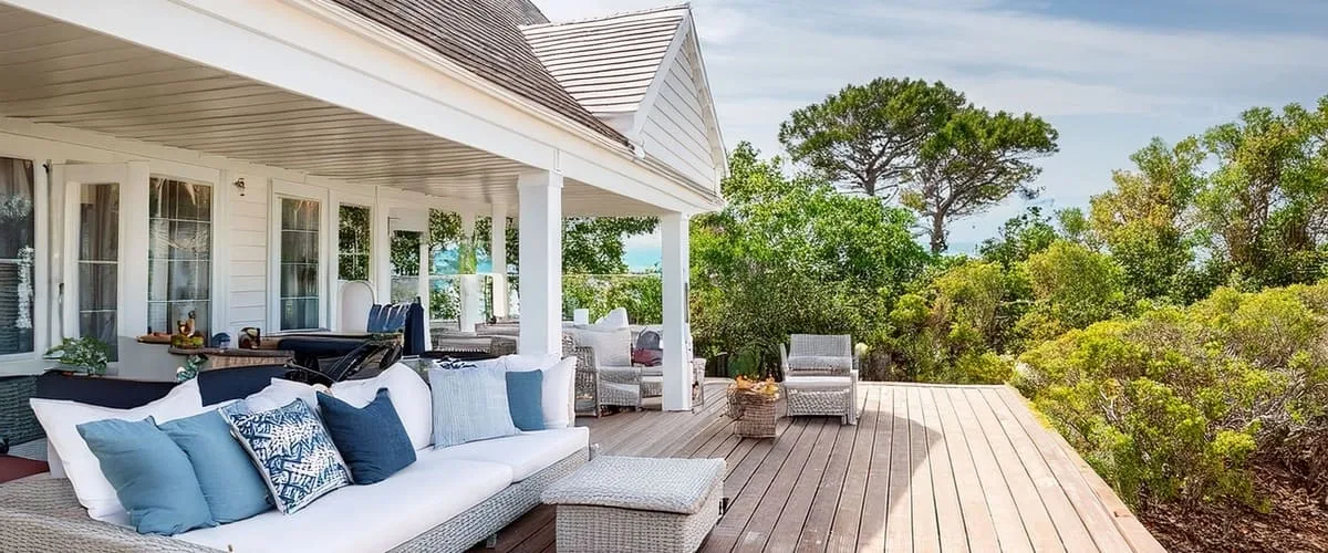 A bright and airy white coastal-style outdoor living room featuring a light grey wicker sofa with blue and white pillows on a wide timber deck overlooking lush green trees and the ocean.