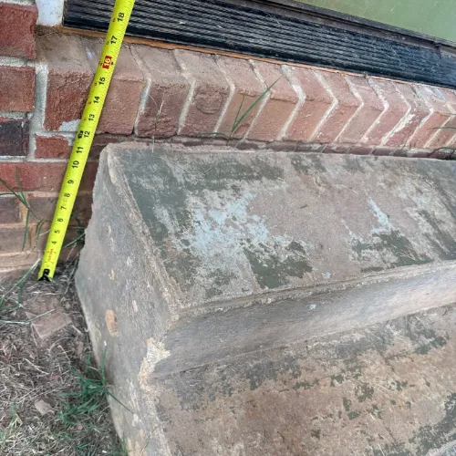 a weathered piece of reclaimed stone or concrete, possibly a window sill or step, next to a brick wall and a measuring tape