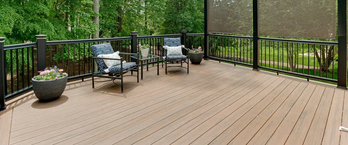 An elevated outdoor living deck featuring dark metal railings, comfortable patio chairs, and decorative planters overlooking a lush green forest.