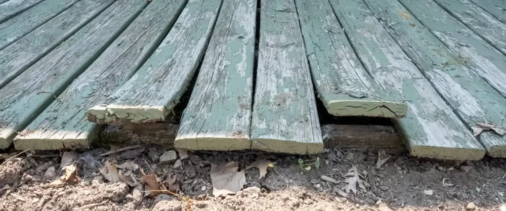 Low-angle view of a warped deck with peeling green paint and rotting wooden boards resting directly on the soil.