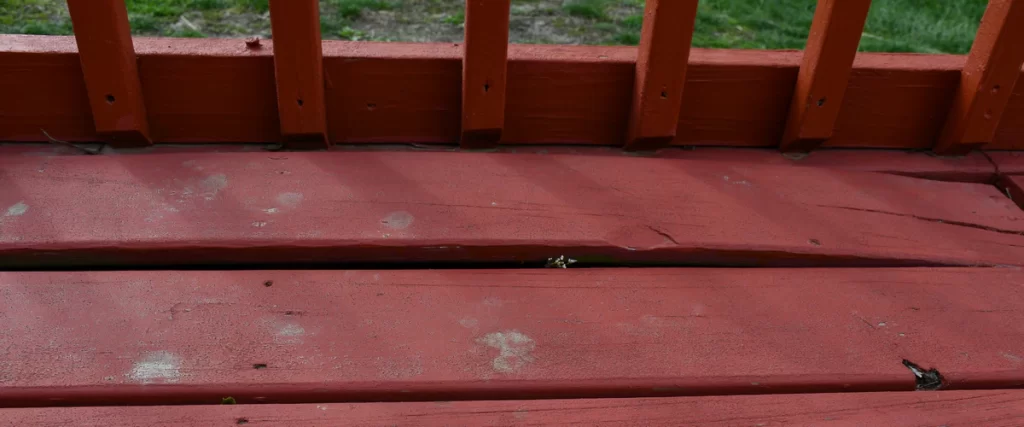 A warped deck made of red painted wood boards showing significant gaps and uneven leveling under a railing.