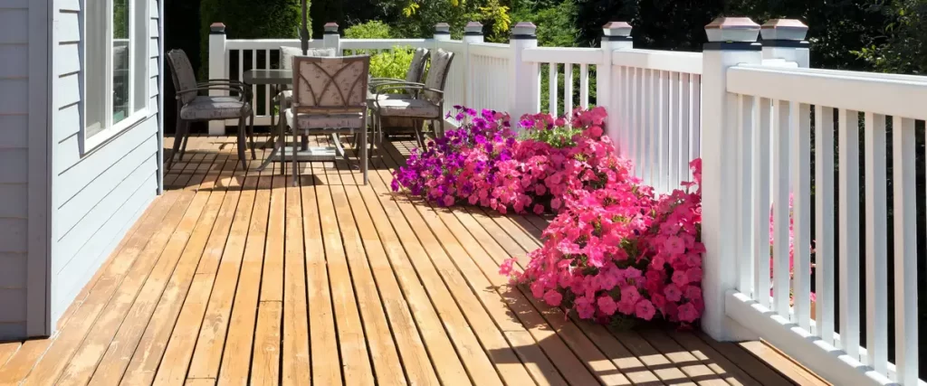 A sun-drenched light wood deck with clean white railings and large, vibrant clusters of pink and magenta petunias in bloom.