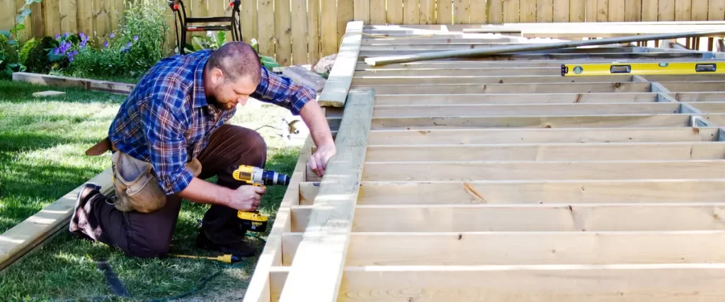 A carpenter in a blue plaid shirt kneeling on a lawn while using a power drill to secure a board to a new wooden deck frame.