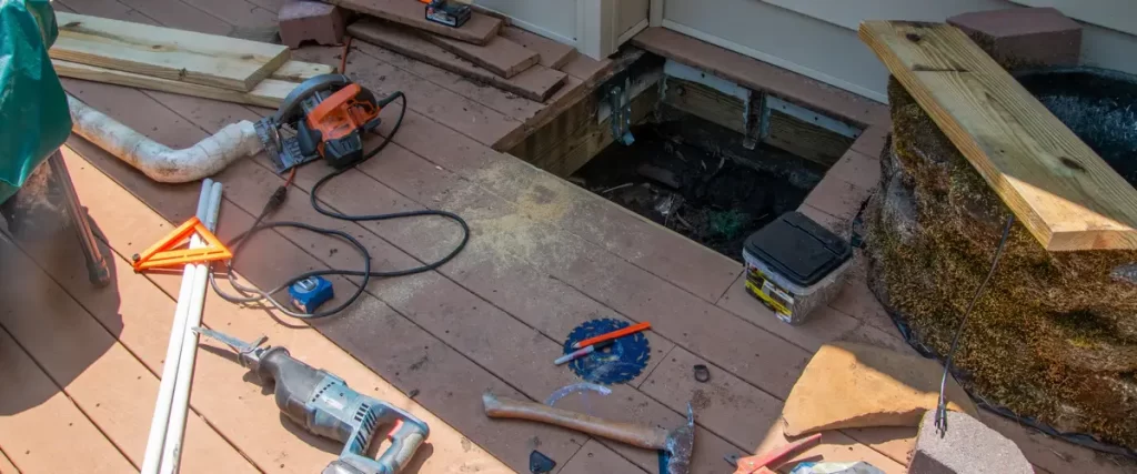 An active deck repair site showing a structural opening in the floorboards surrounded by a circular saw, reciprocating saw, and hand tools.
