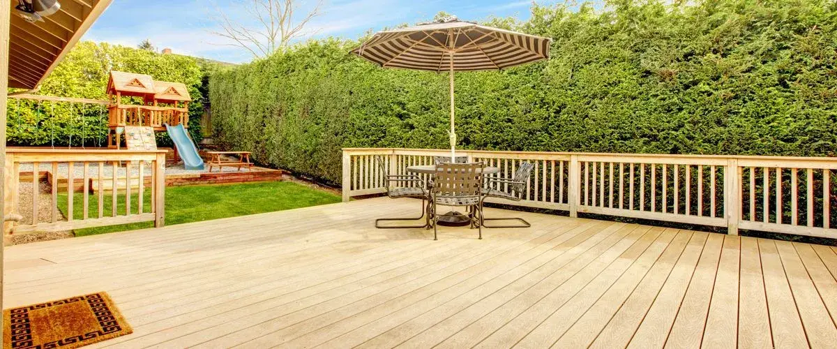 A large, light-colored wooden deck with a striped umbrella dining set, featuring a view of a backyard children's playset and a tall privacy hedge.