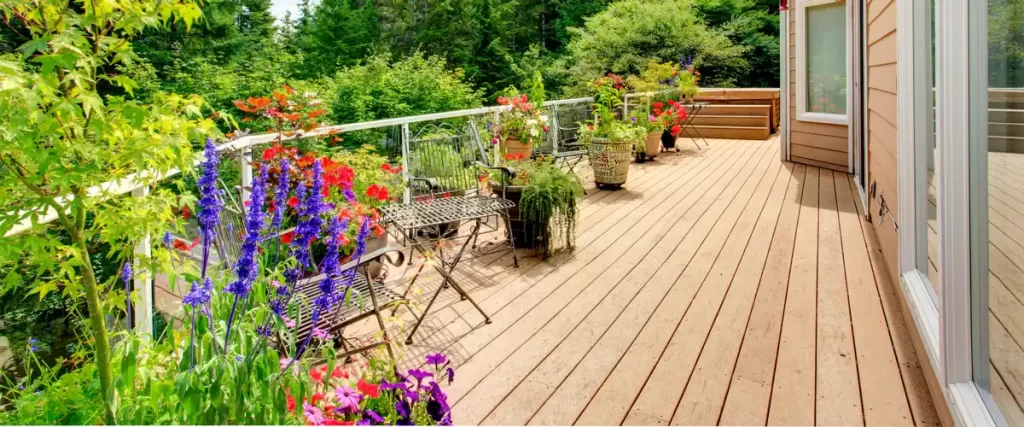 A perspective view of a long wooden deck lined with potted flowers, purple salvia, and lush green trees in the background.