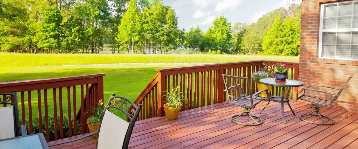 A beautifully stained reddish-brown wooden deck with metal patio furniture and a small bistro table overlooking a lush green lawn.