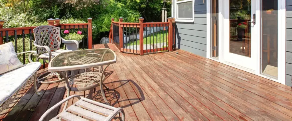 A weathered wooden deck with visible grain wear featuring a glass-top table and metal chairs against a gray house exterior.