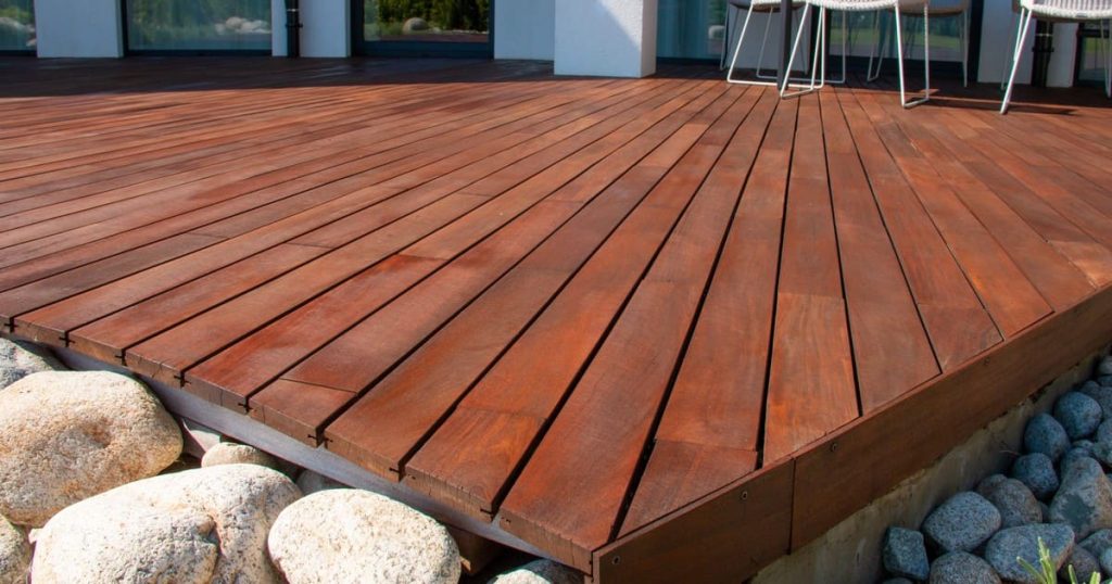 A close-up view of a rich, reddish-brown stained wood deck with smooth river rocks along the edge, illustrating why spring is the best time to repair your deck in Chattanooga.