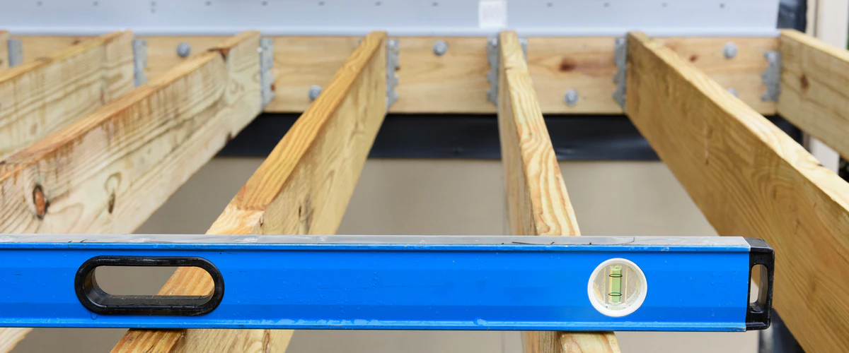 A close-up view of a blue spirit level resting horizontally across wooden framing joists, ensuring an even build where the joists connect to the deck ledger board with metal brackets.