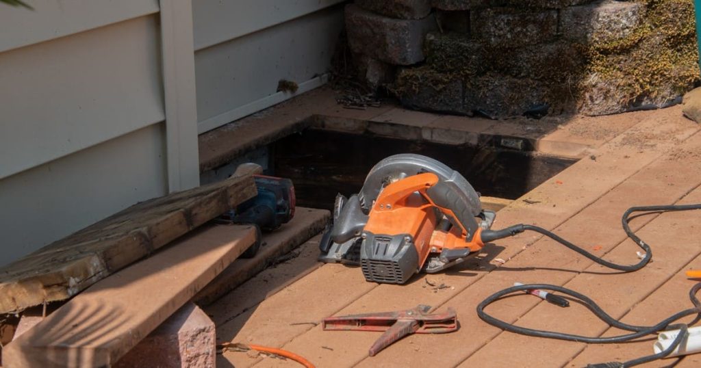 An orange circular saw resting on brown deck boards beside a cutout section near light house siding and a mossy stone planter for under deck drainage systems TN.