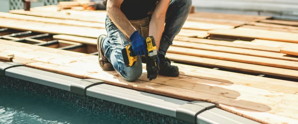 A contractor kneeling and using a yellow power drill to install new wooden boards during a poolside deck replacement project.