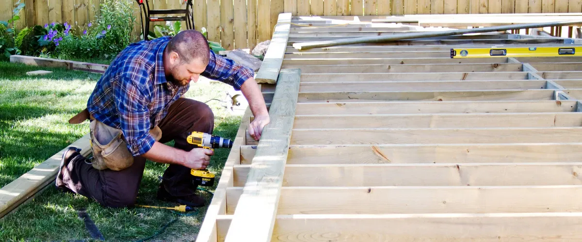 A male contractor kneels on the grass using a yellow power drill to secure the outer rim board to the framing joists as part of a secure deck ledger build.