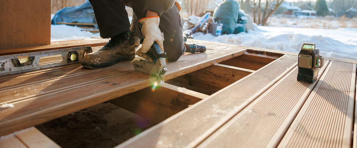 A contractor wearing boots and gloves kneels in the snow, using a specialized fastening tool to replace wooden deck boards over exposed joists alongside a laser level.