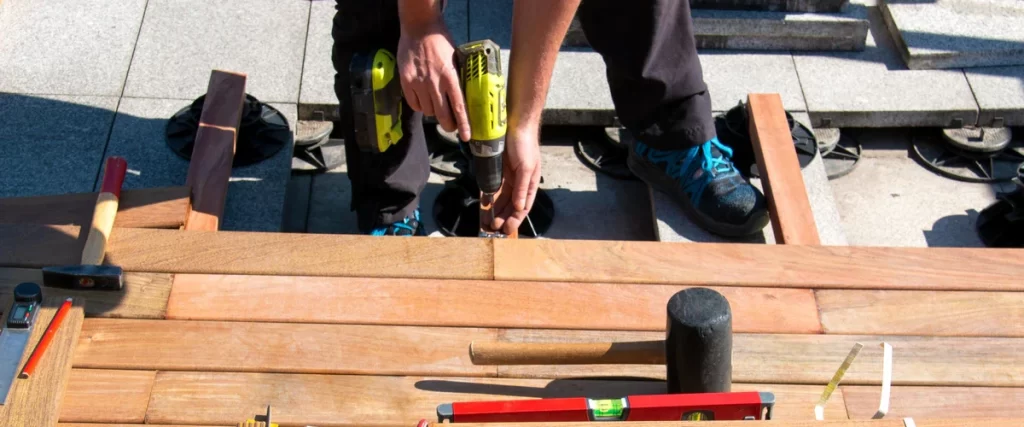 A worker using a power drill to fasten wooden boards over adjustable pedestal supports during an outdoor deck building project, with various tools like a hammer, mallet, and level nearby.