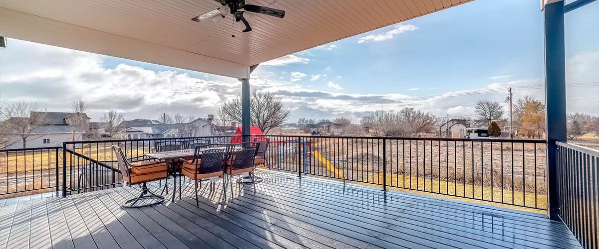 A spacious covered deck painted gray, featuring a ceiling fan, black metal railings, and an outdoor dining set looking out over a residential backyard.