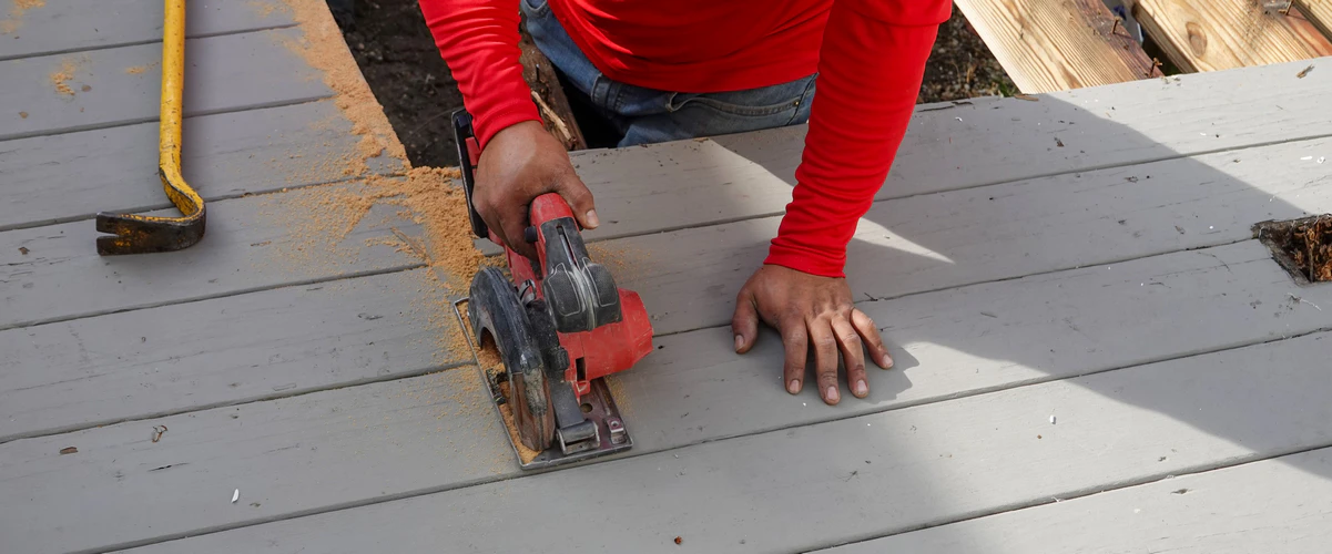 A person in a red shirt using a power circular saw to cut through painted grey wooden deck boards next to a yellow pry bar during a deck repair.