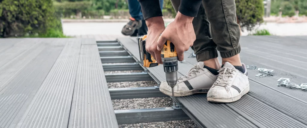 Close-up of a person using a yellow power drill to install grey composite deck boards onto a metal joist frame using hidden fasteners to replace an old surface.