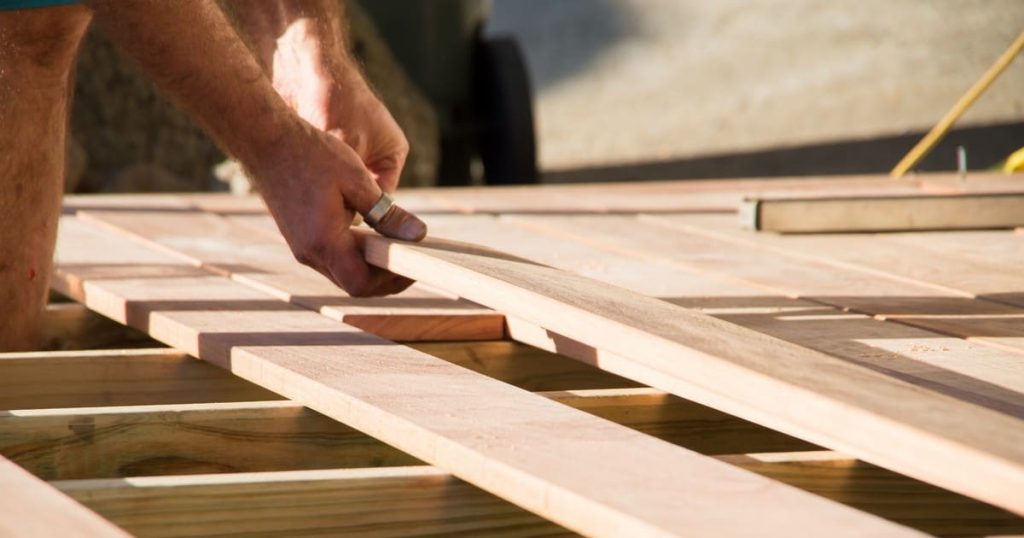 A close-up of a builder's hands carefully placing a new wooden board onto the joist frame, illustrating the manual labor involved when determining how long does it take to build a deck.