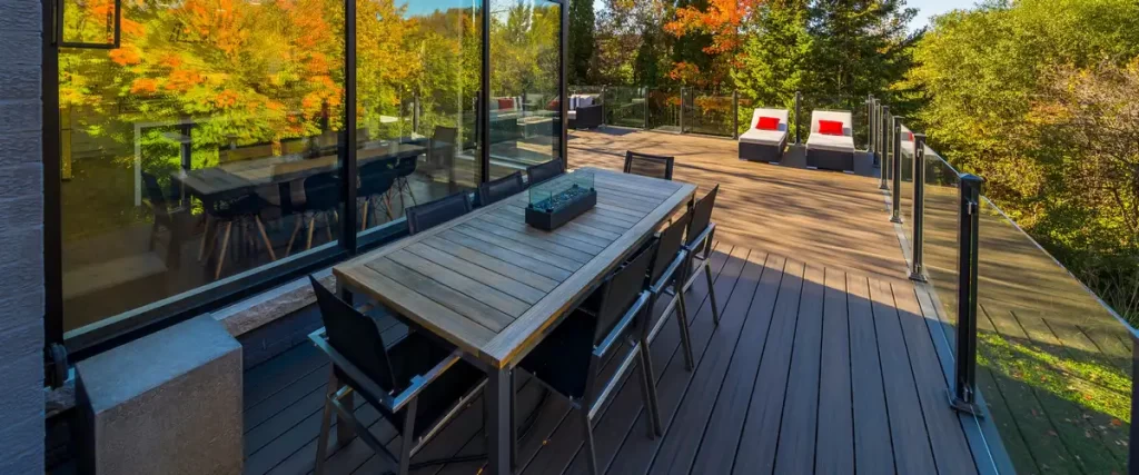 Modern outdoor deck with dark grey decking, featuring a large wooden dining table, a glass fire pit centerpiece, sleek glass railings, and white lounge chairs against a backdrop of autumn trees.
