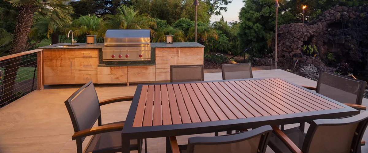 A modern deck area featuring an outdoor kitchen with a built-in stainless steel grill and light wood cabinets, situated next to a slatted dining table and matching furniture.