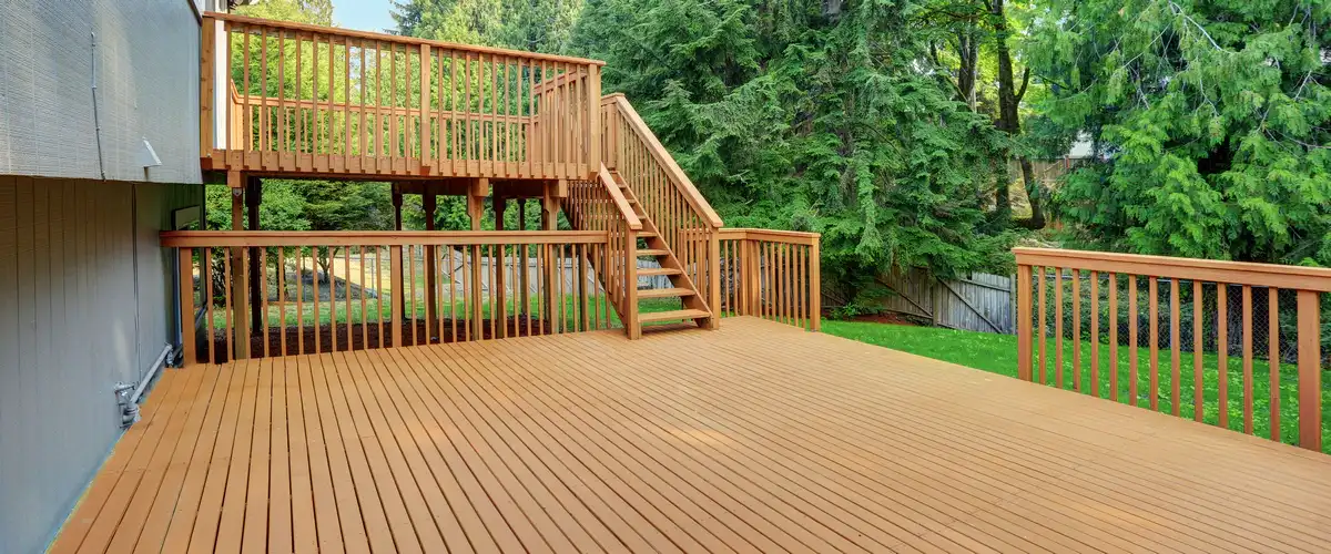 A large multi-level wooden deck attached to a gray house, featuring wooden stairs connecting the expansive lower platform to a smaller upper balcony.