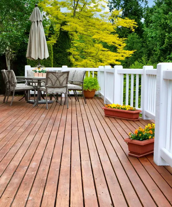 A finished redecking project featuring a wooden surface decorated with a patio dining set, umbrella, white railings, and potted bright flowers against a backdrop of lush green trees.