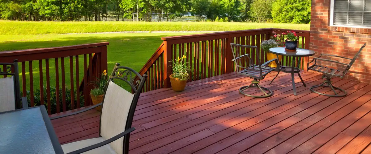 A red painted wooden deck featuring a glass-top table, metal swivel chairs, and bright potted plants overlooking a sprawling green lawn with outdoor furniture.