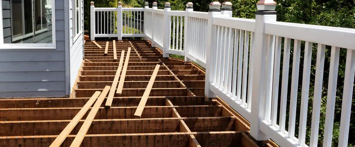 An outdoor deck undergoing repair, featuring exposed brown wooden joists, new support beams laid across, and a white railing system.