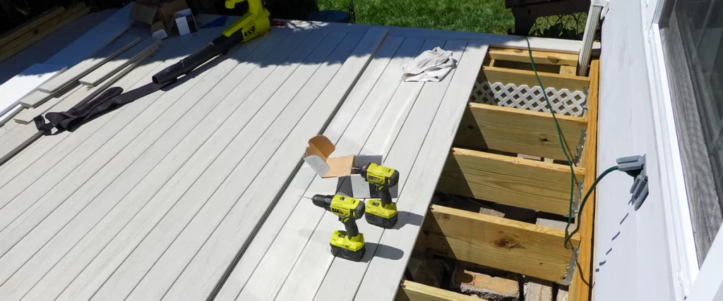 Two yellow power drills resting on light grey deck boards next to exposed wooden joists and a white downspout during a deck drainage and surface renovation.