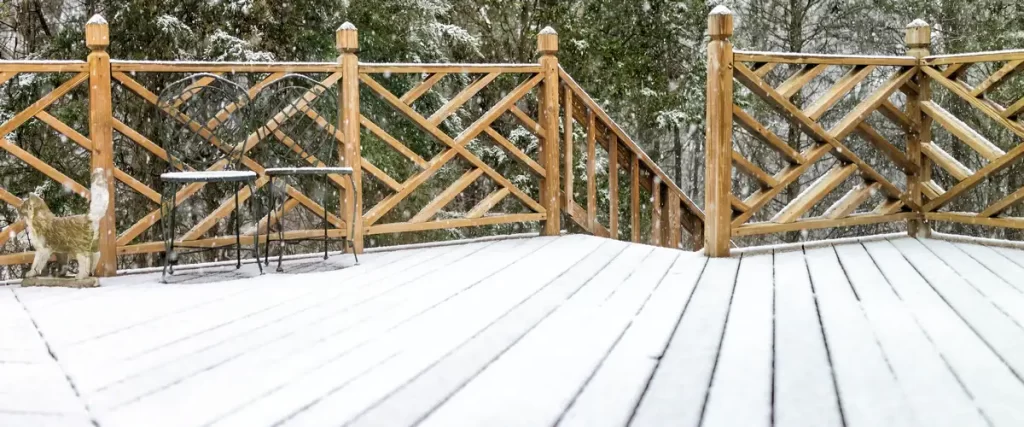 A wooden deck featuring a decorative criss-cross lattice railing, a metal chair, and a cat statue, all covered in a fresh layer of snow during the winter season.