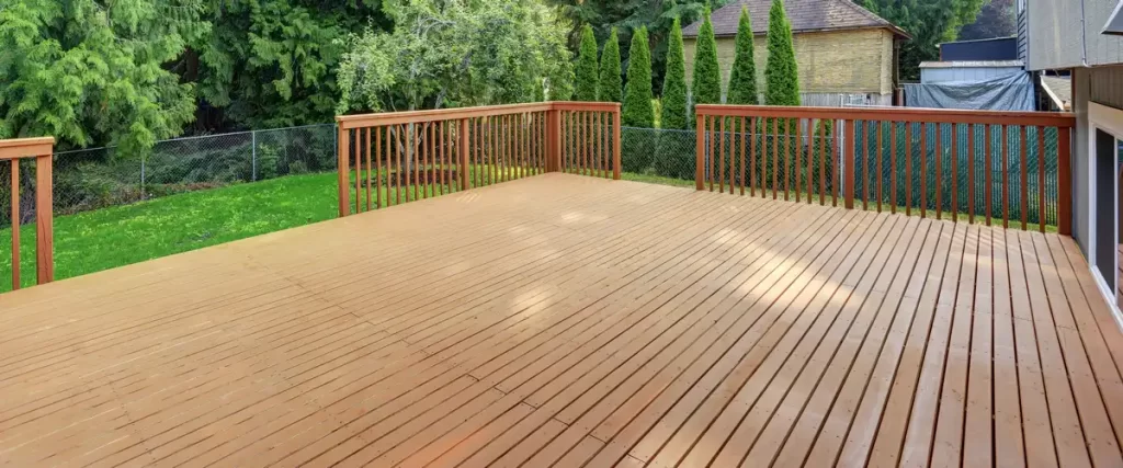 A spacious, empty wooden deck featuring matching wood railings, overlooking a vibrant grassy backyard with tall green privacy trees.