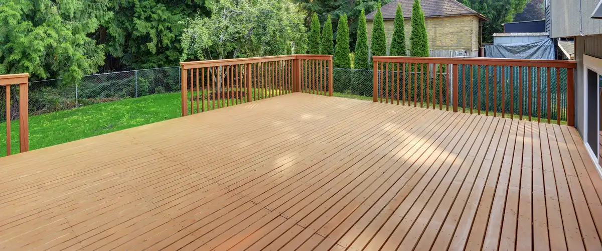 A spacious, empty wooden deck featuring matching wood railings, overlooking a vibrant grassy backyard with tall green privacy trees.