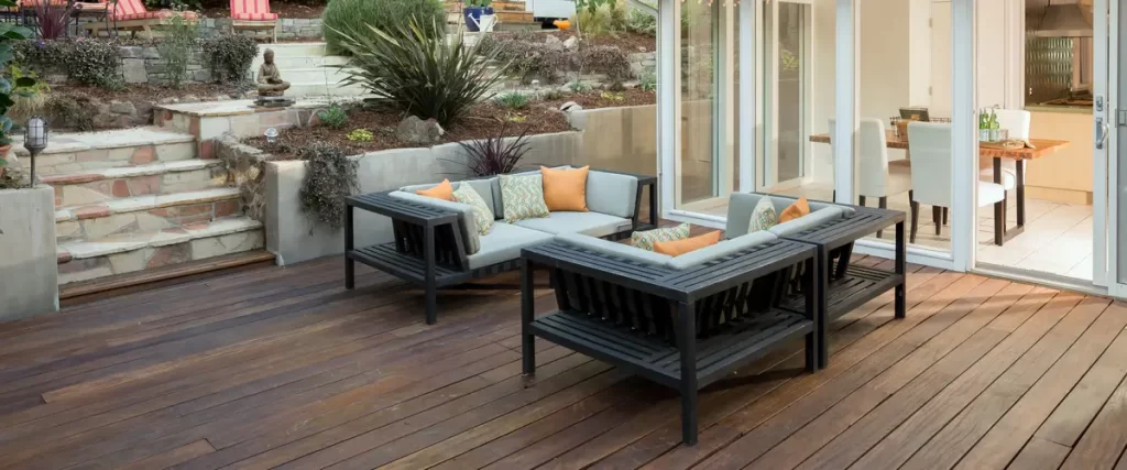 A beautiful backyard wood deck featuring a modern gray outdoor sectional with orange pillows, situated next to landscaped stone steps and large glass patio doors.