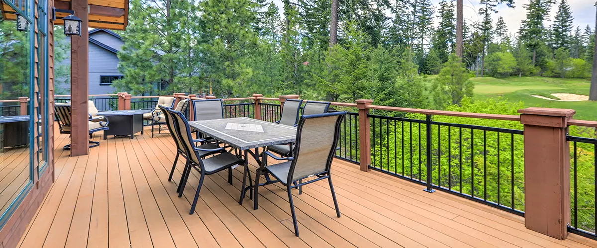 A finished wooden deck featuring a dining table, chairs, fire pit, and black metal railings, overlooking a scenic golf course and pine trees.
