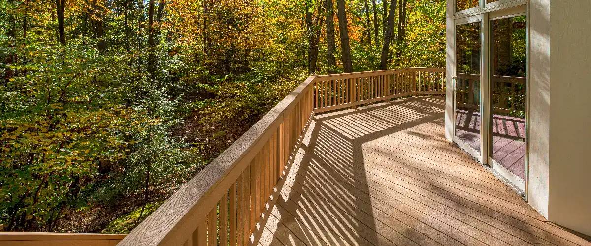 A beautiful curved wooden deck with matching wood railings attached to a home with sliding glass doors, overlooking a scenic forest with autumn foliage.