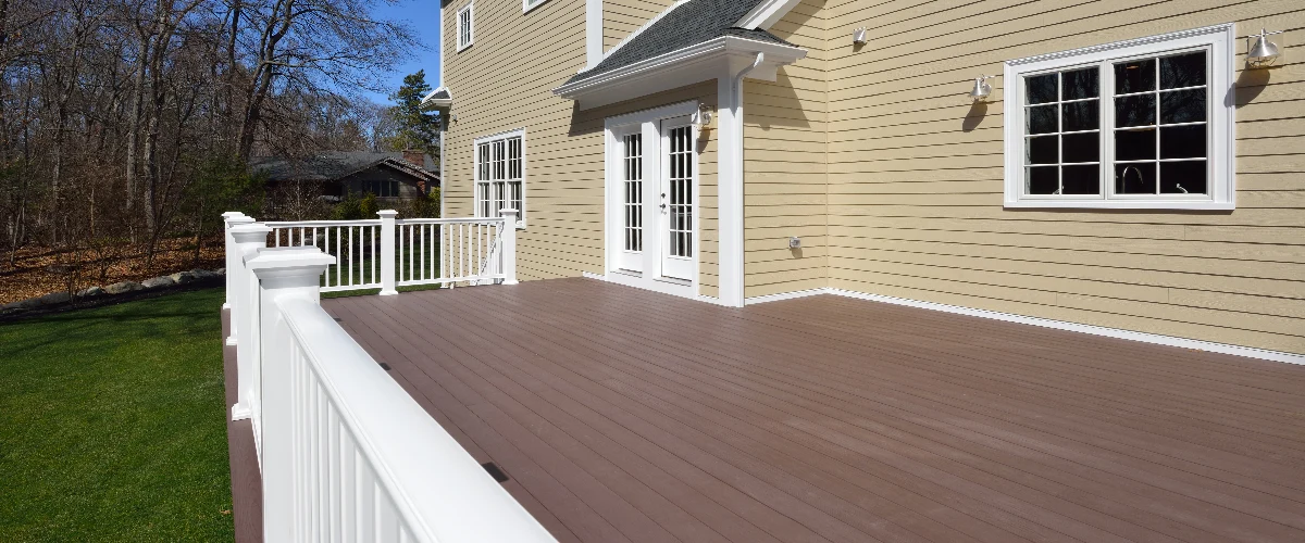 A large, expansive brown deck featuring clean white railings, attached to a yellow home with white trim and classic French doors leading to the backyard.