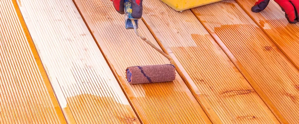 A person wearing red work gloves uses a small paint roller to apply a protective honey-colored wood stain to grooved timber decking boards, showing the contrast between the treated and untreated wood.